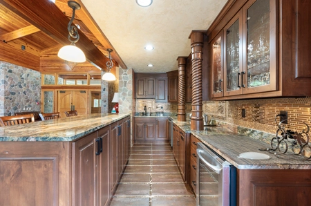A kitchen space with bar seating, a sink, oven, dishwasher, glass cabinets, dark tiled backsplash, and stone flooring