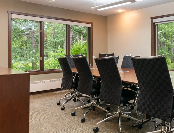 A meeting room with a conference table, wheeled chairs, and two large windows looking out into the surrounding greenery